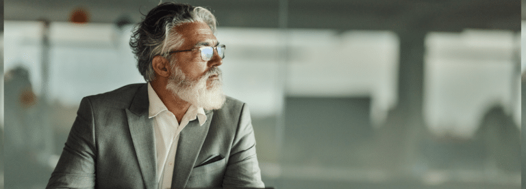 A businessman with white hair and a white beard, wearing a gray suit, looks pensively into the distance.