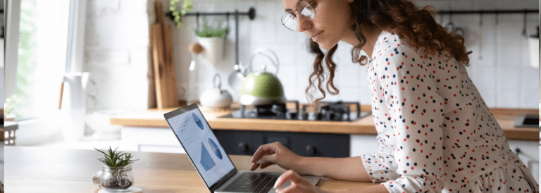 A woman using a laptop computer. She has long, curly brown hair, and she is wearing wireframe glasses with a polka dot dress.