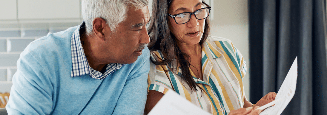 A man and a woman looking at papers. The man has short white hair and he is wearing a blue sweater, and the woman has long dark hair and a pinstriped button-up shirt.