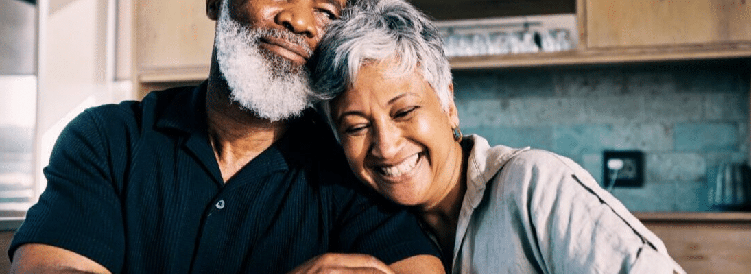 An elderly man and woman smile and laugh together, while leaning against each other affectionately. They both have white hair, and are sitting in a kitchen.