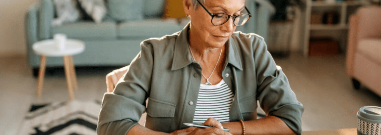 A woman writing in a notebook. She has short white hair and round glasses, and she is wearing a striped t-shirt and olive green overshirt.