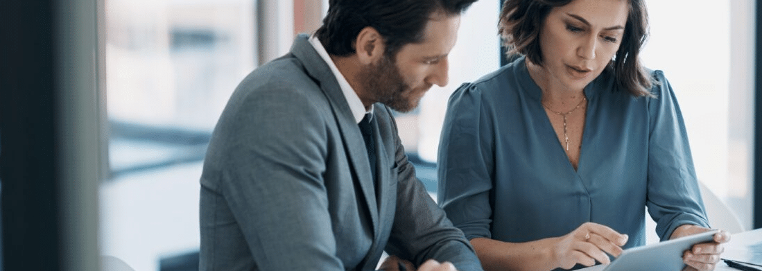 A man wearing a gray suit is looking at a tablet held by a woman in a blue dress.