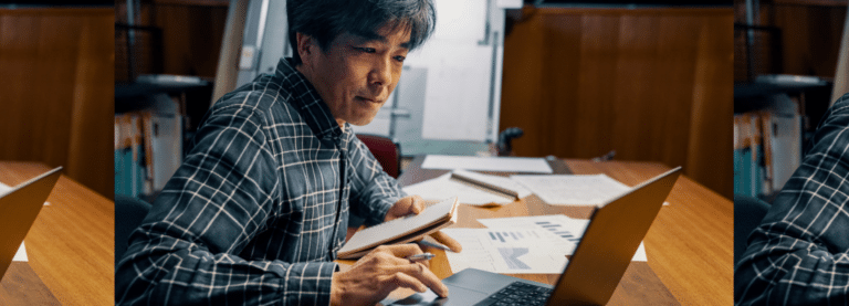 A man wearing a plaid shirt sits at a tabletop covered in documents. He is holding a pen and a notebook, and using a laptop computer.