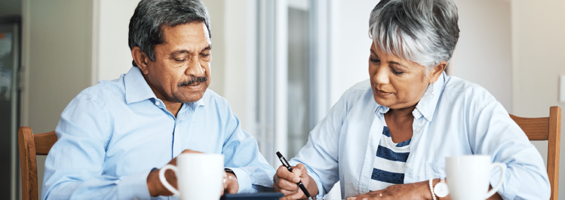 An older couple sitting at the kitchen table. They are both wearing button-up blue shirts. Both are looking at a calculator, and they are revising a document with an ink pen.