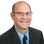 A headshot of associate James Barnes. He is wearing wireframe eyeglasses and a black suit.