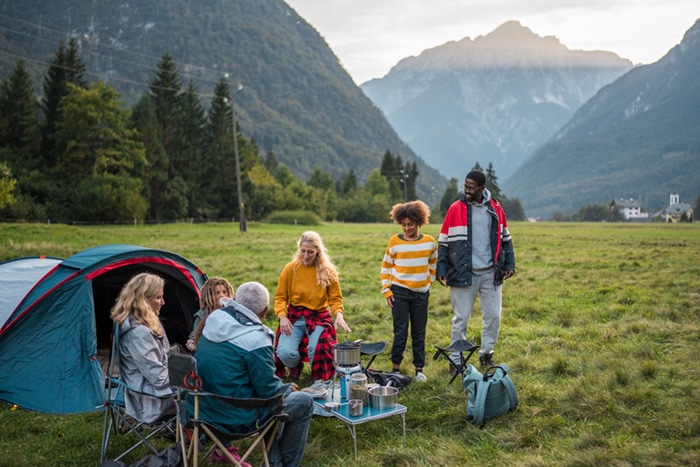 A diverse family engages in conversation outside a tent, surrounded by mountains.